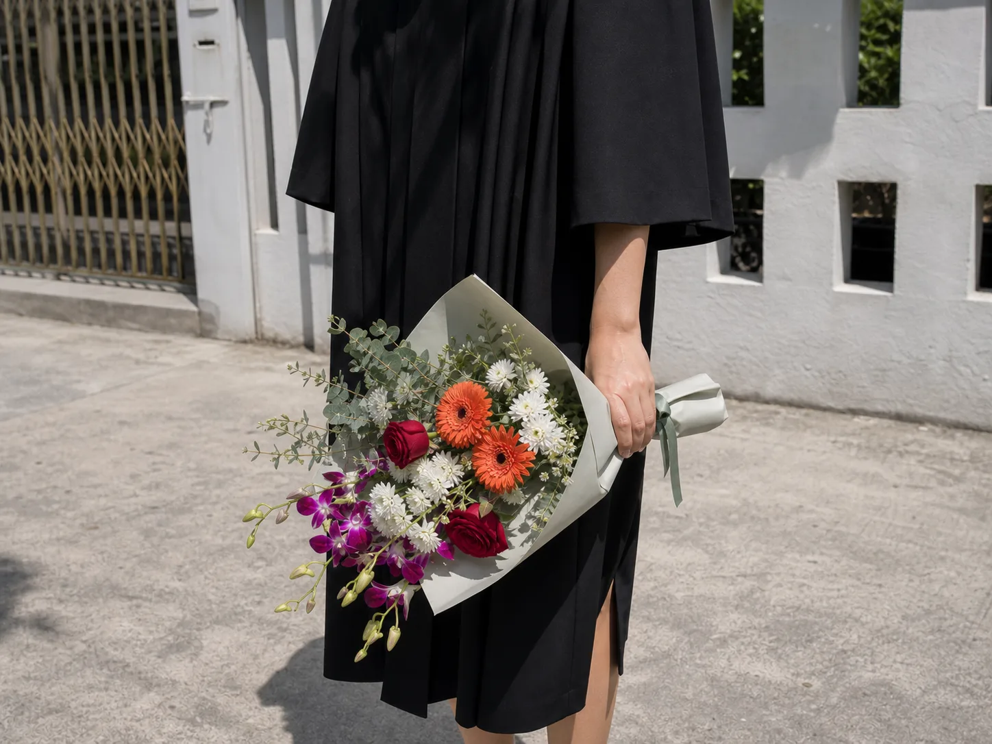 A graduation posy with a wrapped handle held one-handed at a graduate&rsquo;s side in bright outdoor sunlight